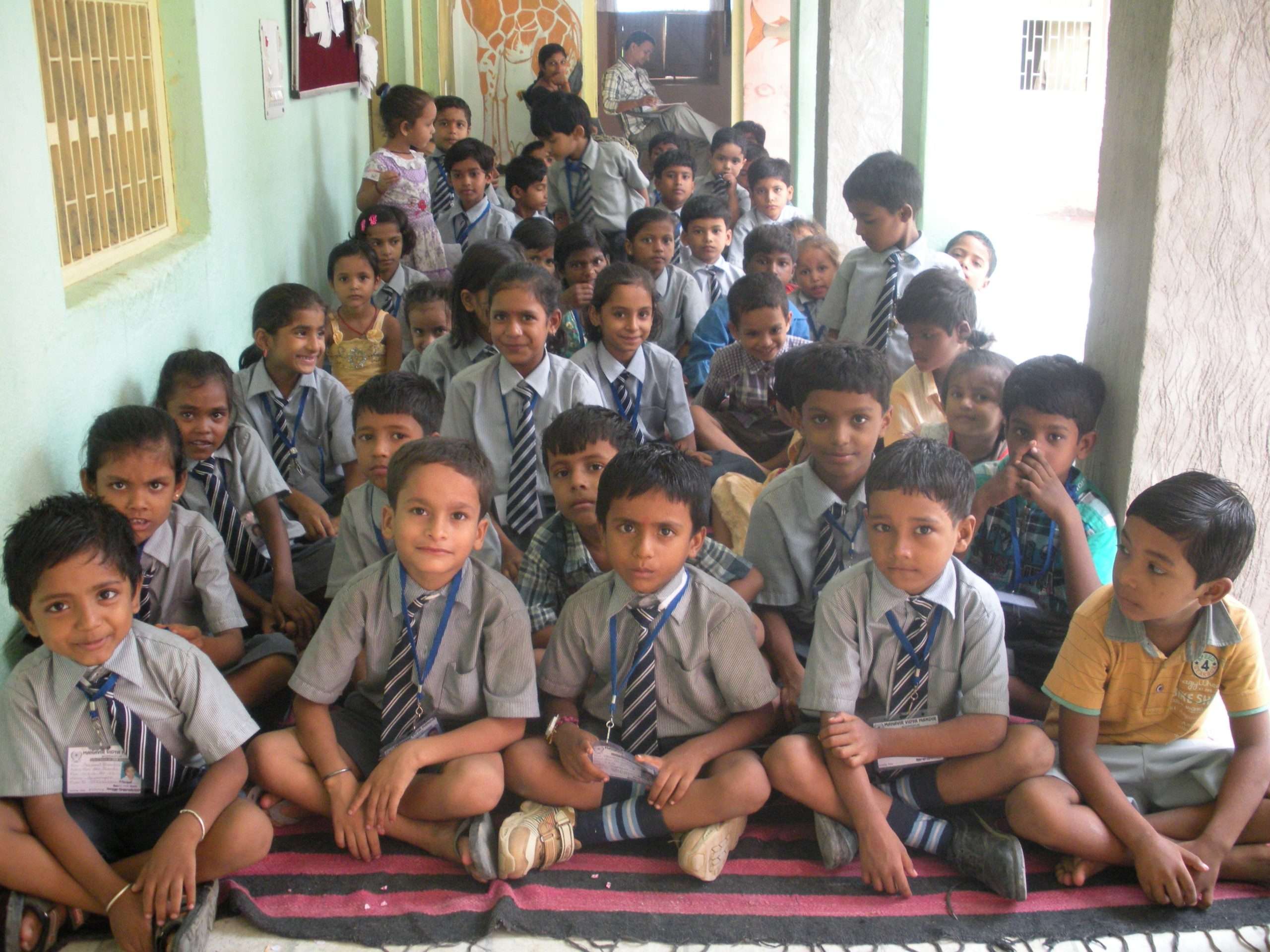 Many children in school uniform seated cross-legged in the school verandah