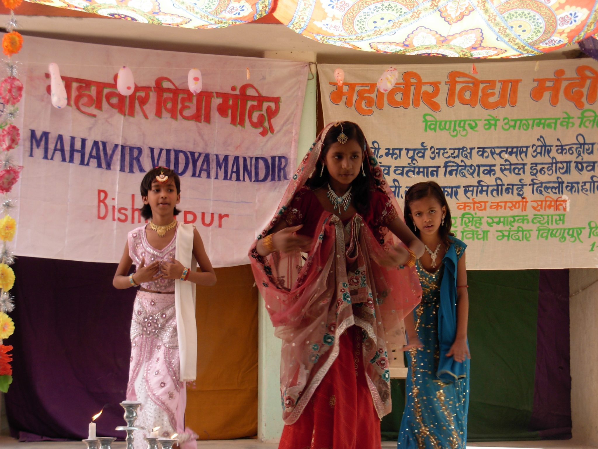 Three girls in traditional costumes performing a classical dance on stage at Mahavir Vidya Mandir