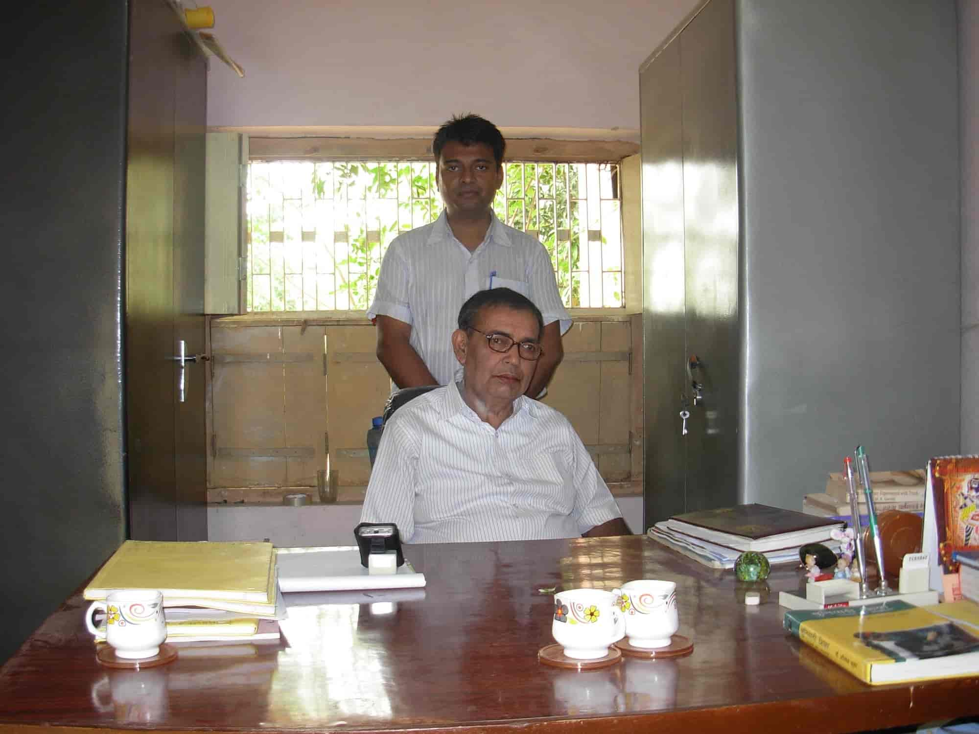 A senior Trust member seated at his wooden office desk with files and tea cups, younger associate standing behind