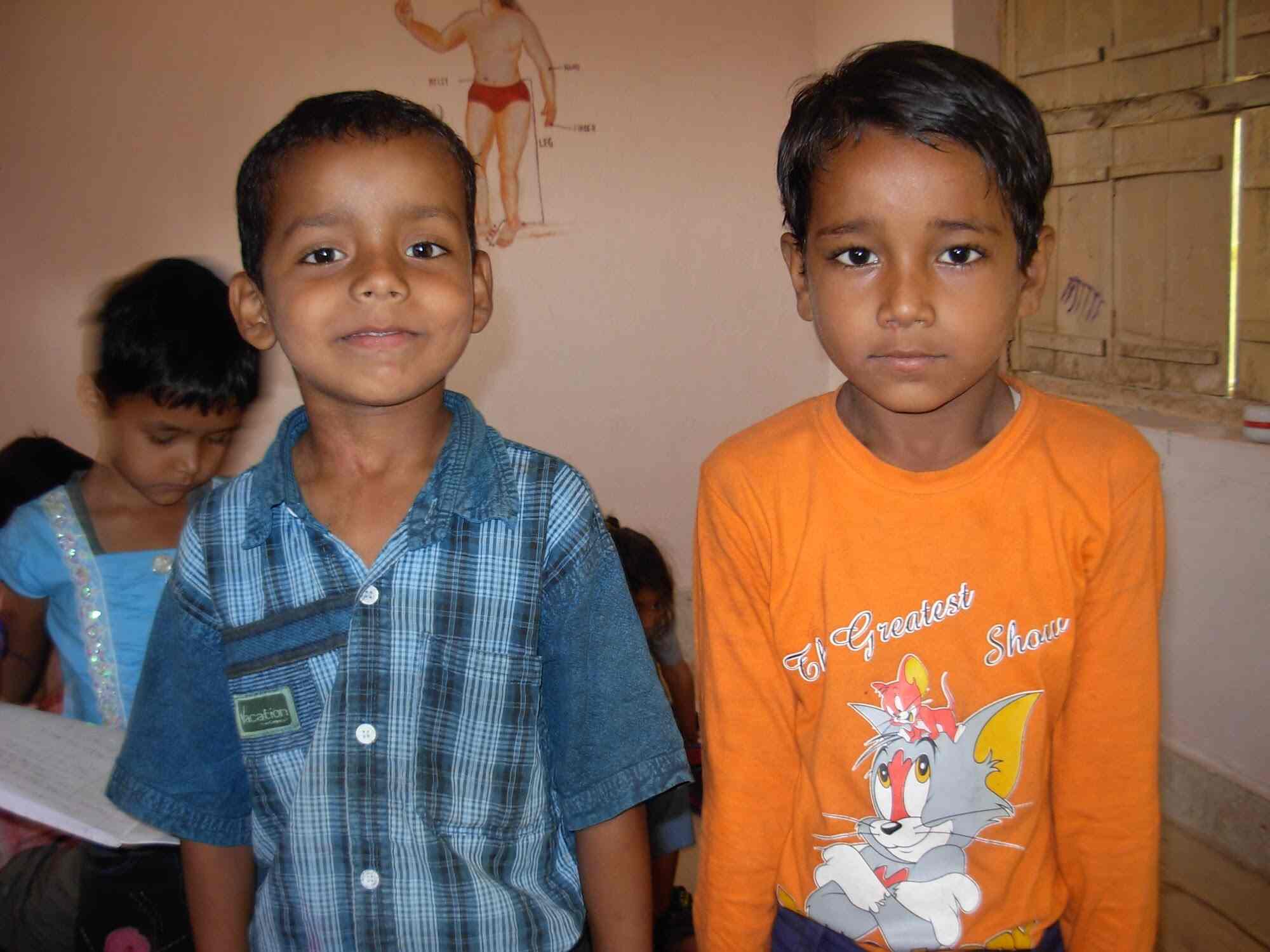 Two boys in casual clothes posing in a classroom, one in a blue check shirt, one in orange t-shirt