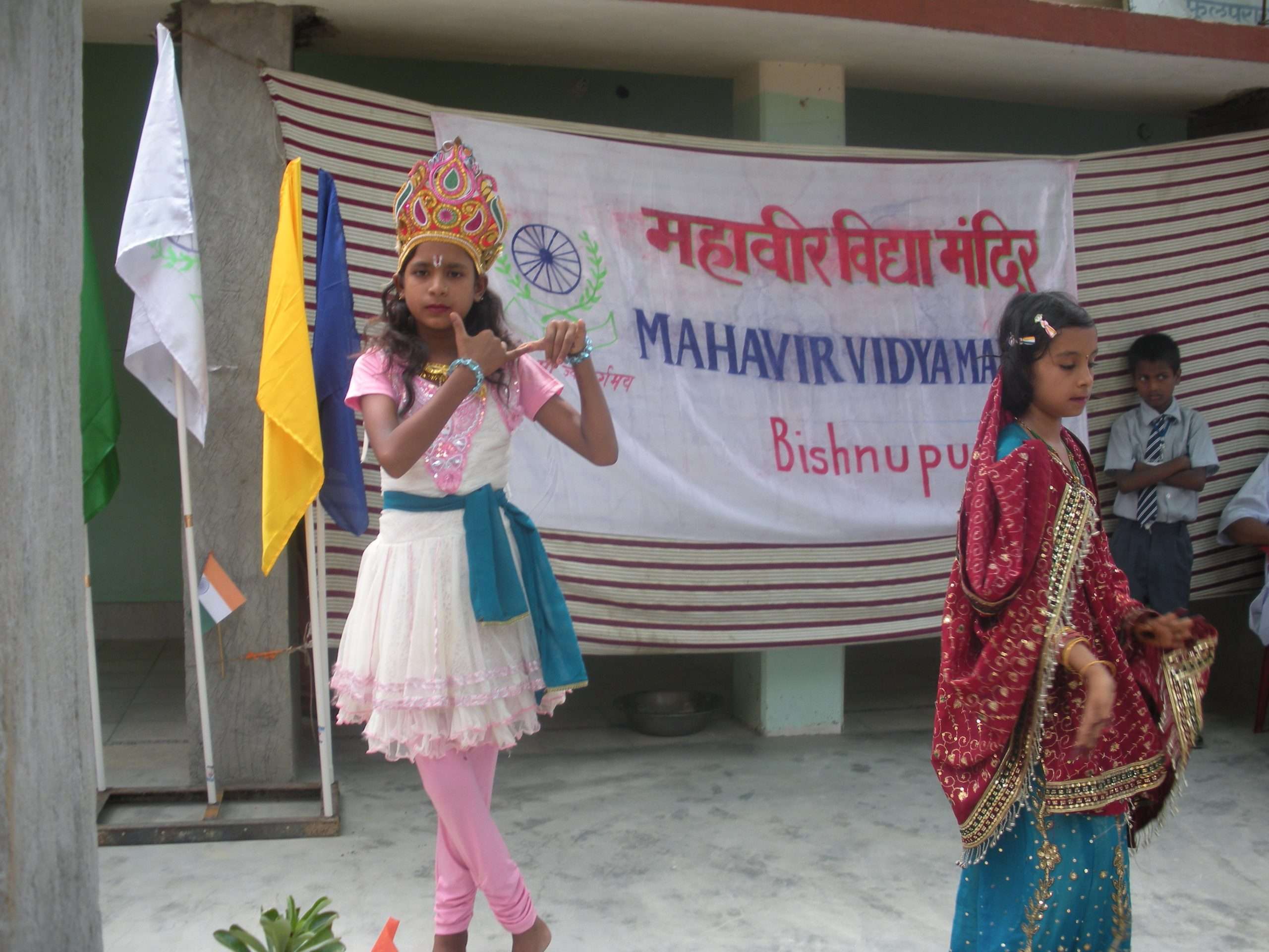 Young girl in Krishna costume with crown and flute, performing on stage with another dancer